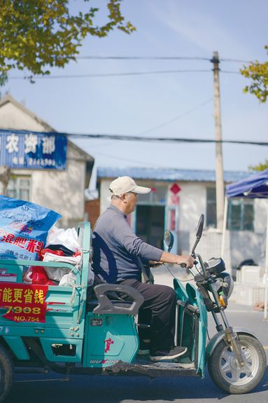 An older villager drives a load of product on his turquoise three wheeler in the city