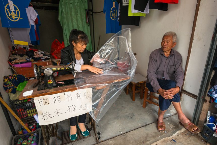 A tailor repairing a raincoat for a local farmer in Tuocheng Town, Guangdong province. From mending clothes, umbrellas to suitcases, the tailor shop offers a wide range of services. (2016)