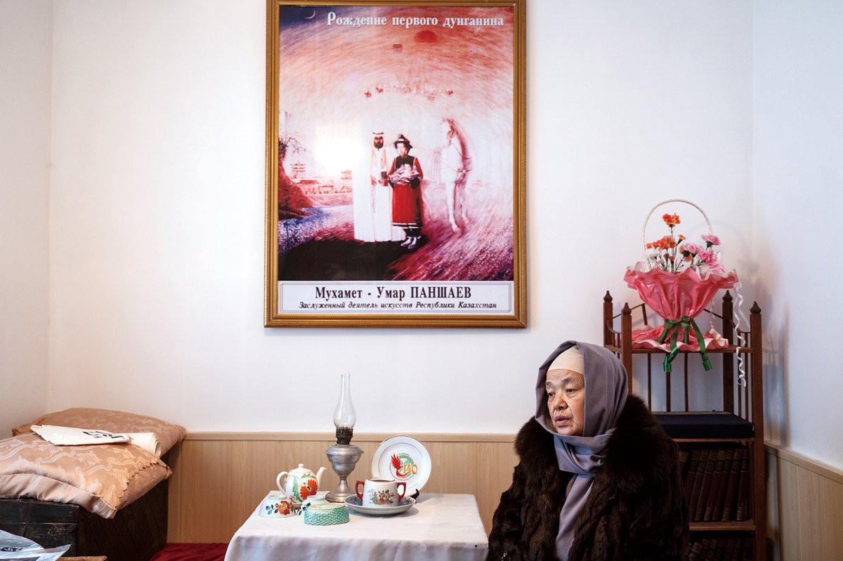 A Dungan elder sits in front of a painting depicting the origin story of Hui people, connecting the origins of Dungan people