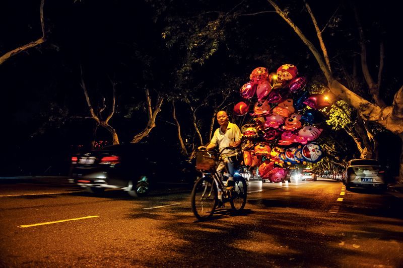 An elderly man rides a bike around the nightclub gates to sell his balloons to young couples looking to impress